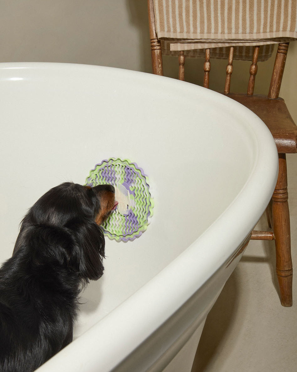 Dog playing with a lick mat inside a bathtub next to a wooden chair.